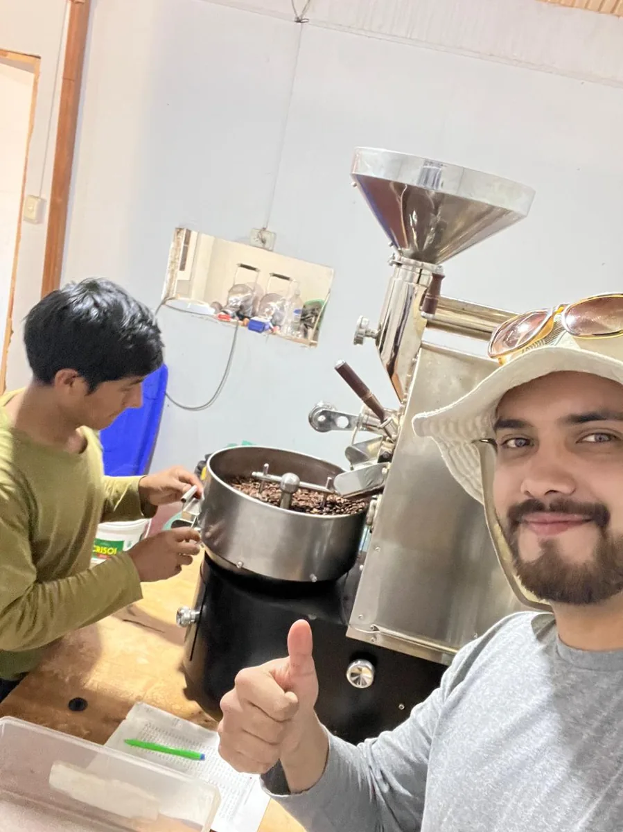 Adrian sonríe junto a un tostador de café mientras otra persona revisa el tueste